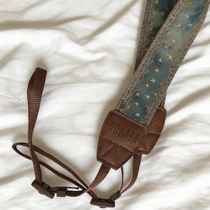 May include: A camera strap with a blue and gold star pattern, brown leather accents, and the brand name "ADHARA" embossed on a leather patch. The strap has a brown herringbone border and brown nylon connectors.