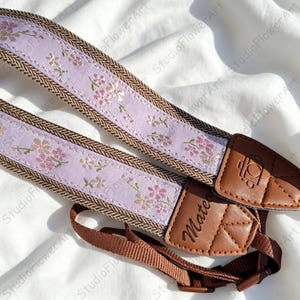 May include: A camera strap featuring a light purple fabric band with a repeating pink and green floral pattern, bordered by brown herringbone webbing. The strap includes brown leather end tabs with a camera icon and the name Mateo embossed.