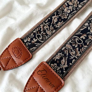 May include: A camera strap featuring a black and tan floral woven pattern with brown leather ends. One leather end is embossed with a camera icon, while the other is personalised with the name Eliana in a dark script font.