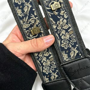May include: Two camera straps with a navy blue floral pattern and gold accents. Each strap has a small, gold-coloured camera emblem. The straps are held by a person. The word "Sophia" is printed on the black leather end.