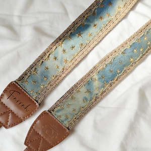 Peut inclure: Deux sangles d'appareil photo en tissu bleu effet jean, ornées de broderies d'étoiles dorées. Les extrémités en cuir marron portent le nom "Farrah" et une icône d'appareil photo. Les bords sont bordés d'un motif en chevrons marron.