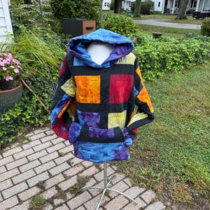May include: A long-sleeve hooded sweatshirt featuring a patchwork design of black fabric panels interspersed with tie-dye squares in shades of orange, red, yellow, purple, and blue. The garment includes a front kangaroo pocket and a hood.