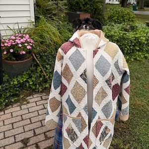 May include: A long-sleeved hooded jacket featuring a quilted diamond pattern in various colours including maroon, blue, tan, and cream. The garment has two front patch pockets and a light-coloured trim along the hood and front opening edges.