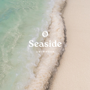 May include: A close-up of a wave breaking on a sandy beach. The water is clear and turquoise, and the sand is white. The text "Seaside Swimwear" is superimposed on the image.