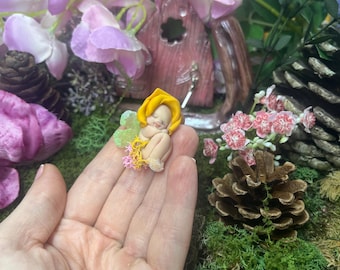 May include: A miniature fairy figurine with a yellow bonnet and delicate wings, nestled in a bed of green moss. The fairy is surrounded by miniature flowers, a small wooden house, and pinecones, creating a whimsical fairy garden scene.