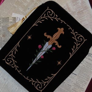 May include: Black velvet pouch with an embroidered design featuring a dagger, roses, and ornate gold-colored trim. The pouch has a gold tassel and is set against a backdrop of vintage newspaper pages.