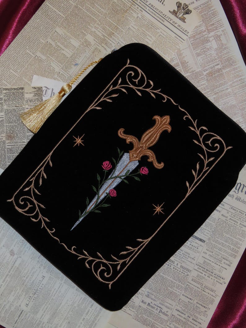 May include: Black velvet pouch with an embroidered design featuring a dagger, roses, and ornate gold-colored trim. The pouch has a gold tassel and is set against a backdrop of vintage newspaper pages.