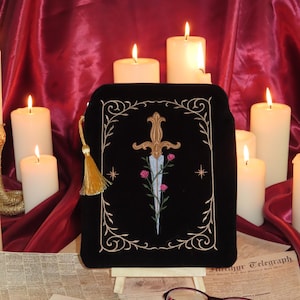 May include: Black velvet pouch with gold embroidered design of a dagger, roses, and decorative border. The pouch has a gold tassel zipper pull. Lit candles and vintage newspapers are in the background.