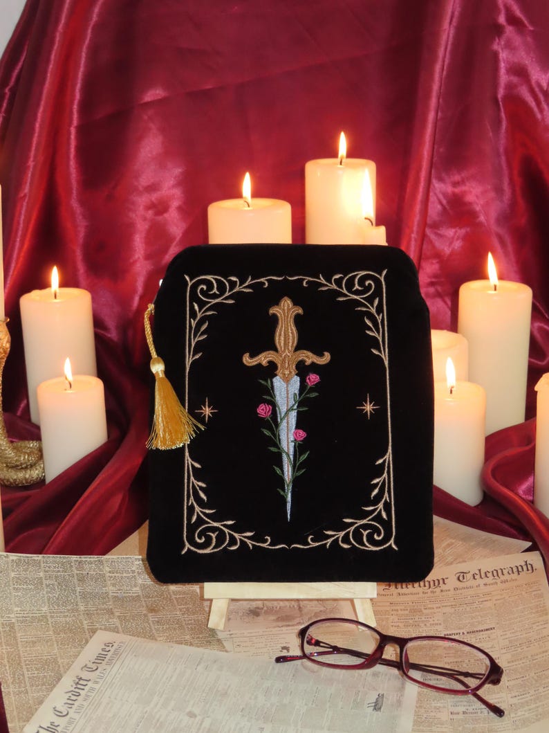 May include: Black velvet pouch with gold embroidered design of a dagger, roses, and decorative border. The pouch has a gold tassel zipper pull. Lit candles and vintage newspapers are in the background.