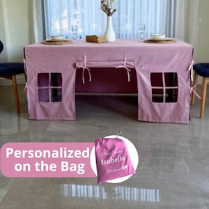 Puede incluir: Una funda de mesa rosa con forma de casa de juegos, con ventanas y lazos. La mesa está puesta con platos y un jarrón con flores. En primer plano, una bolsa rosa con el texto "Merry Christmas Isabella We love you".