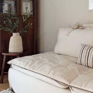 Puede incluir: Escena de salón de tonos neutros con un sofá color crema con una parte superior acolchada y un cojín a rayas. Un jarrón tejido con vegetación se encuentra sobre una pequeña mesa de madera. Un mueble de madera oscura con libros y una foto enmarcada está al fondo.