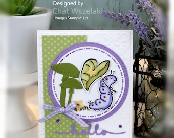 May include: A handmade greeting card featuring a white embossed floral background, a green polka dot panel, and a purple circular frame. The card displays a purple caterpillar, green mushrooms, a yellow butterfly, and a purple ribbon bow with the text hello.