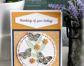 May include: A handmade greeting card featuring a mustard yellow panel with the text "thinking of you today" in a white hexagon. Below, a white circular die-cut overlay displays three butterflies, light green leaves, and small yellow flowers.