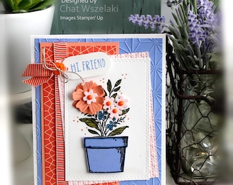 May include: A handmade greeting card featuring a blue pot with flowers, layered on orange and blue geometric patterned paper. The card includes a white tag with the text HI. FRIEND, a striped ribbon, and a stitched white rectangular panel.