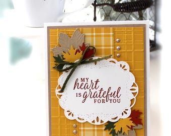 May include: A rectangular greeting card featuring a yellow textured background with a plaid pattern strip. It includes die-cut paper maple leaves in red, yellow, and green, a dark green twine bow, and a white scalloped label with the text My heart is grateful for you.