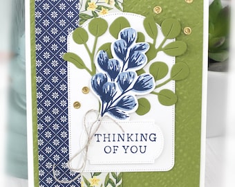 May include: A handmade greeting card featuring a textured olive green background, a navy blue floral patterned strip, and a white die-cut panel. It includes green paper eucalyptus sprigs, a blue stamped botanical element, twine, and the text THINKING OF YOU.
