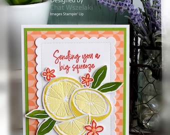 May include: A rectangular greeting card featuring a peach and white geometric pattern background, a white scalloped border, and the text "Sending you a big squeeze" in red. The card is decorated with yellow lemon slices, green leaves, and small red flowers.