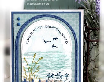 May include: A rectangular greeting card featuring an arched die-cut window. The scene depicts a beach with sand, tall grass, and birds. The text reads WISHING YOU SUNSHINE & HAPPINESS. The card is framed in blue with two small blue circular embellishments.