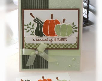 May include: A handmade greeting card and a matching tag featuring stamped images of three pumpkins in dark green, orange, and light green. The card includes a light green embossed background, a dark green striped panel, and the text a harvest of BLESSINGS.