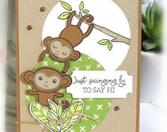 May include: A handmade greeting card featuring two brown monkeys, one hanging from a tree branch and one below, with a green patterned circle background. The card includes the text Just swinging by TO SAY HI and is accented with small brown beads.