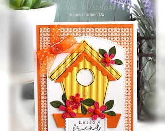 May include: A rectangular greeting card featuring a yellow corrugated birdhouse with a scalloped orange roof, decorated with pink flowers, green leaves, and an orange polka-dot ribbon bow. The card displays the text "hello friend" in black script.