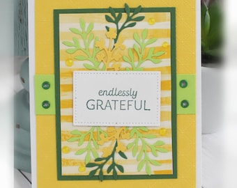 May include: A rectangular greeting card with a yellow textured background featuring horizontal white stripes. The card is decorated with dark green, light green, and yellow die-cut foliage, small yellow gems, and a white label with the text endlessly GRATEFUL.
