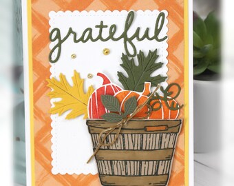 May include: A handmade greeting card featuring a brown basket filled with orange pumpkins and green leaves. The word grateful is die-cut in dark green above the basket. The background is a light orange plaid pattern with a white scalloped border.