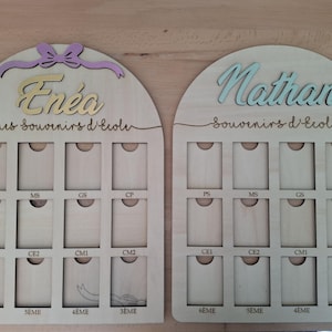 Peut inclure: Deux tableaux de souvenirs scolaires en bois. L'un est intitulé "Enéa" avec un nœud violet et des lettres dorées, l'autre "Nathan" en bleu. Chaque tableau comporte des emplacements pour les souvenirs scolaires, avec les niveaux scolaires.