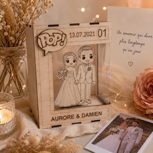 Può includere: Una scatola espositiva in legno con un'illustrazione tagliata al laser di una sposa e uno sposo. La parte superiore include il testo POP!, 13.07.2021 e 01. La base è incisa con AURORE & DAMIEN. La scatola ha un pannello frontale trasparente.