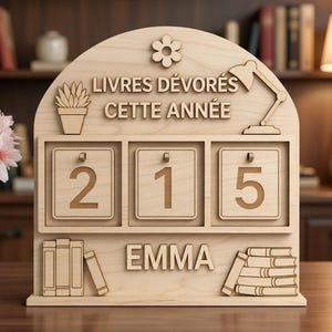 Peut inclure: Présentoir en bois en forme d'étagère avec le texte "LIVRES DÉVORÉS CETTE ANNÉE" et le nom "EMMA". Il comporte une fleur, une plante en pot, une lampe de bureau et les chiffres 215. Le présentoir est en bois clair.