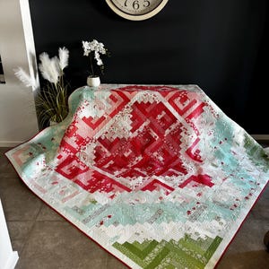 May include: A red, white and green quilt with a geometric pattern. The quilt is folded in half and is sitting on a table in front of a black wall.
