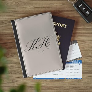 May include: A gray passport holder with black stitching and the initials "K.H." in a cursive font. The holder is lying on top of a blue passport with the word "SPORT" printed on the cover. There are also two airline boarding passes visible in the image.