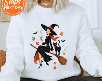 May include: A white long-sleeve sweatshirt featuring a graphic illustration of a witch in a black dress and pointed hat sitting on a broomstick, holding a green drink, accompanied by a black cat, orange and green autumn leaves, and bats.