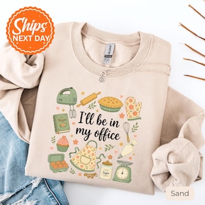 May include: A sand-colored crewneck sweatshirt featuring a graphic print of baking tools including a mixer, rolling pin, pie, oven mitt, recipe book, cupcake, apron, sugar jar, scale, and piping bag with the text I'll be in my office.