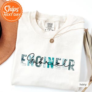 May include: An ivory short-sleeve t-shirt featuring the word ENGINEER in a distressed teal and white block font, with the name Gabriel written in a black cursive script overlaying the center of the text. A gold necklace rests on the collar.
