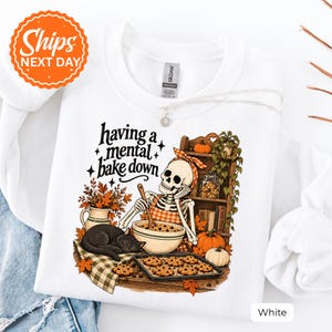 May include: A white crewneck sweatshirt featuring a graphic of a skeleton wearing an orange checkered apron and headscarf, mixing cookie dough in a bowl. A black cat sleeps nearby, with text reading having a mental bake down in black script.