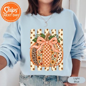 May include: A light blue crewneck sweatshirt featuring a central graphic of a pumpkin decorated with leopard print, gingham, and floral patterns. The pumpkin is topped with a peach-colored bow and sits against a background of vertical polka dot stripes.