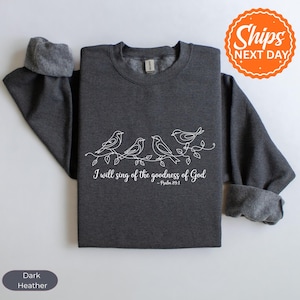 May include: Dark heather gray sweatshirt with white line art of birds on a branch. The text "I will sing of the goodness of God - Psalm 89:1" is printed below the birds. The sweatshirt has a crew neck and rolled-up sleeves.