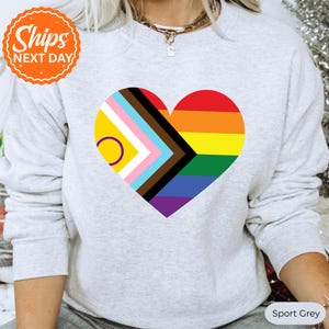 May include: A light gray crewneck sweatshirt featuring a large heart-shaped graphic on the chest. The heart design incorporates the intersex pride flag colors on the left and the traditional rainbow pride flag colors on the right side.