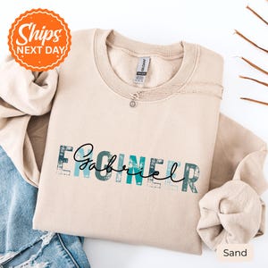 May include: A sand-colored crewneck sweatshirt featuring the word ENGINEER in a distressed teal block font, with the name Gabriel written in a black cursive script overlaying the text. The garment has a Gildan brand label at the neck.
