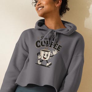May include: A charcoal gray cropped hoodie featuring a graphic of a cartoon coffee mug with arms and legs, waving and walking. The text above the mug reads "DRINK COFFEE" in a bold, vintage-style font. The hoodie has long sleeves and a drawstring hood.