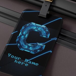 Peut inclure: Une étiquette de bagage rectangulaire noire avec un graphisme numérique bleu en forme de lettre C et des lignes lumineuses rappelant des circuits. Le texte pixélisé Your Name here est inscrit en bleu clair sur la valise marron foncé.