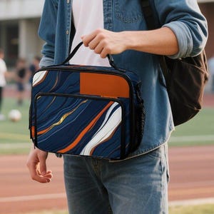 Peut inclure: Un sac à déjeuner rectangulaire présentant un motif de marbre abstrait bleu foncé, orange et blanc avec une poignée supérieure et une poche frontale zippée. Le sac a un design structuré avec un panneau rectangulaire orange uni sur le côté droit.