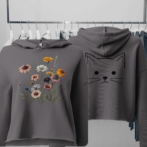 Peut inclure: Un sweat à capuche court gris anthracite orné d'un bouquet de fleurs sauvages colorées sur le devant et d'un dessin au trait noir minimaliste représentant une tête de chat au dos. Le vêtement a des manches longues et un ourlet brut.