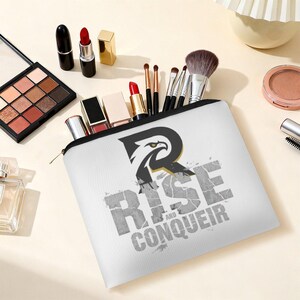 Peut inclure: Une trousse blanche à fermeture éclair ornée d'un motif d'aigle noir et doré intégré dans une grande lettre R, avec le texte RISE AND CONQUER imprimé en gris usé. La trousse contient divers pinceaux de maquillage et rouges à lèvres.