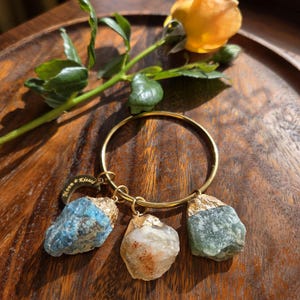 Puede incluir: Una pulsera de aro de metal dorado con tres dijes de piedras preciosas en bruto con tapas doradas. Una piedra es azul, otra blanca con motas naranjas y otra verde. Un pequeño dije de media luna dorada tiene grabado el texto Moon Kissed.