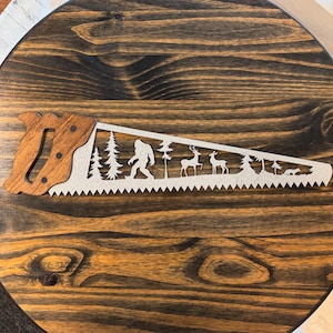 May include: A wooden saw blade with a brown handle. The blade is cut out with a design of a Bigfoot walking through a forest with deer and a fox.