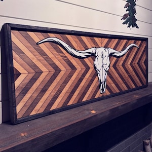 May include: Wooden wall art featuring a white longhorn skull over a chevron pattern. The artwork is framed in black and has a rustic, natural aesthetic. The background is a light-colored wall.