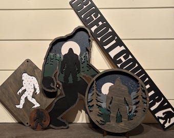 May include: Three wooden wall decorations featuring a Bigfoot silhouette with a forest background. The largest piece says "Bigfoot Country" in black lettering.