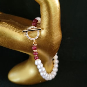May include: A white and red pearl bracelet with a gold toggle clasp. The clasp is decorated with clear rhinestones.